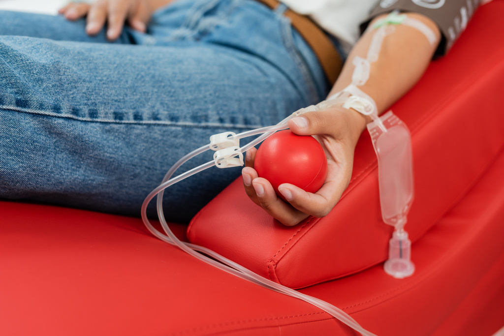 vista parziale di donna multirazziale con set trasfusione tenendo palla di gomma mentre seduto su comoda sedia medica durante la donazione di sangue in laboratorio - Foto, immagini
