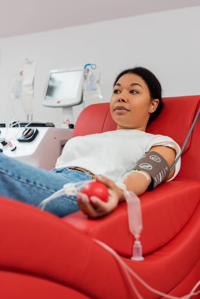многорасовая женщина с набором для переливания крови и медицинским резиновым мячом сидит на удобном медицинском стуле возле автоматизированного оборудования и сдает кровь в лаборатории - Фото, изображение
