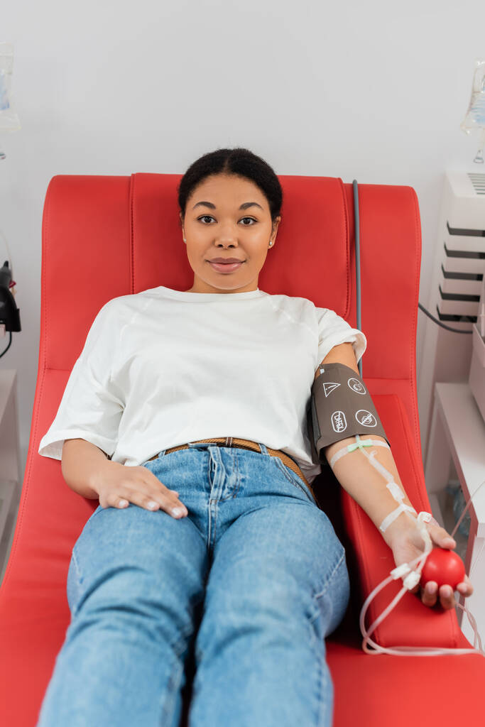 mujer multirracial positiva con manguito de presión, equipo de transfusión y pelota de goma médica sentado en la silla médica y mirando a la cámara durante la donación de sangre en la clínica - Foto, Imagen