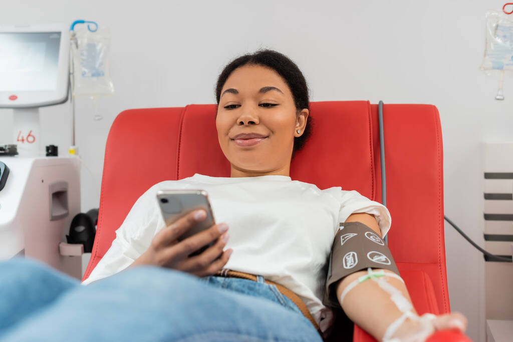 erfreut multirassische Frau in Blutdruckmanschette Nachrichten auf dem Handy, während sie auf dem medizinischen Stuhl in der Nähe der automatischen Transfusionsmaschine in modernen Labor, medizinische Verfahren sitzen  - Foto, Bild