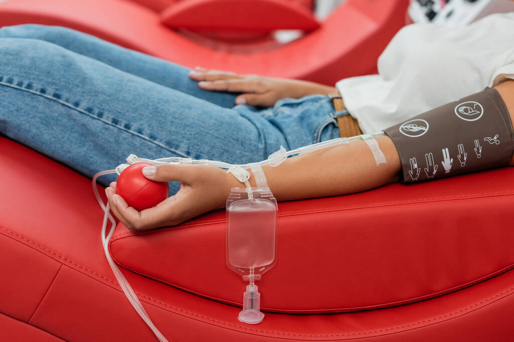 partial view of multiracial woman with transfusion set and medical rubber ball sitting on ergonomic medical chair during blood donation in laboratory - Photo, Image
