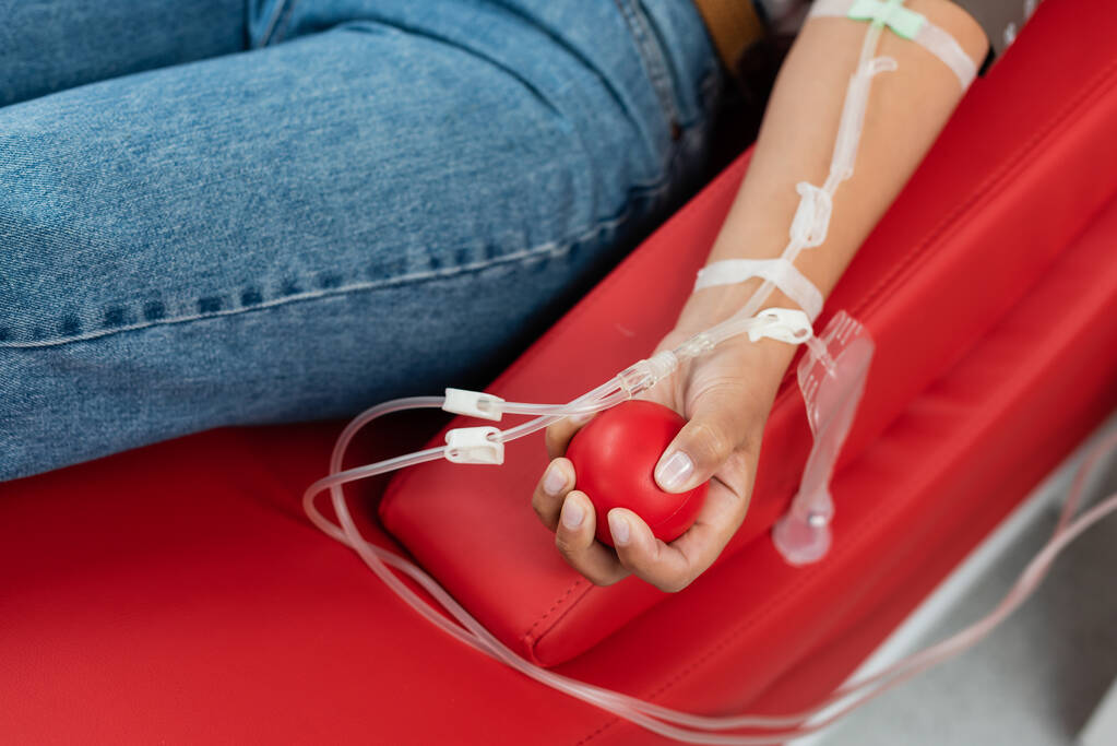 częściowy widok wielorasowej kobiety z zestawem do transfuzji, trzymającej gumową kulę podczas siedzenia na ergonomicznym krześle medycznym podczas oddawania krwi w klinice, zabieg medyczny  - Zdjęcie, obraz