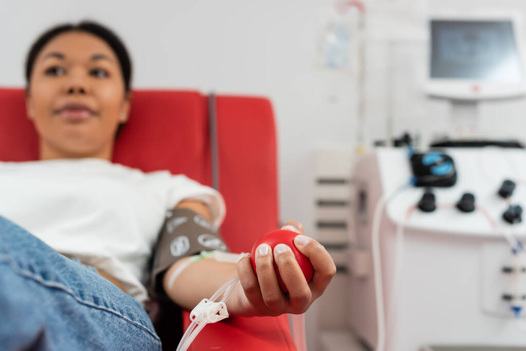 selektiver Fokus des medizinischen Gummiballs in der Hand einer multirassischen Frau, die auf einem bequemen medizinischen Stuhl in der Nähe einer Transfusionsmaschine sitzt, während sie Blut im Labor spendet, verschwommener Hintergrund - Foto, Bild