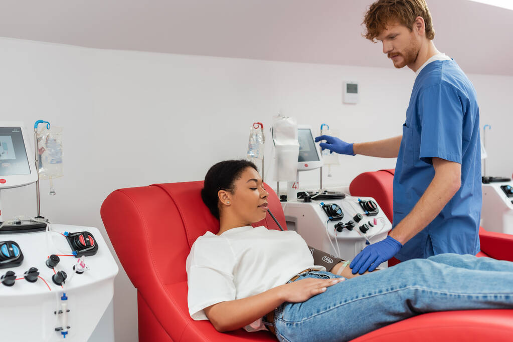 rothaarige Ärztin in blauer Uniform und Latex-Handschuhen, die Transfusionsmaschine in der Nähe einer multirassischen Frau mit Druckmanschette bedient, die auf einem medizinischen Stuhl im Blutspendezentrum sitzt - Foto, Bild
