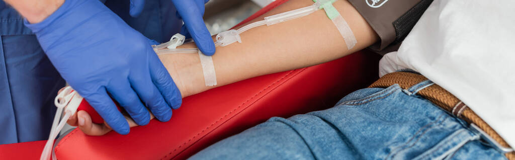 częściowy widok pracownika służby zdrowia w rękawiczkach lateksowych przyklejających plaster do ramienia wielorasowej kobiety siedzącej na krześle medycznym z zestawem do transfuzji krwi, zabieg medyczny, baner - Zdjęcie, obraz