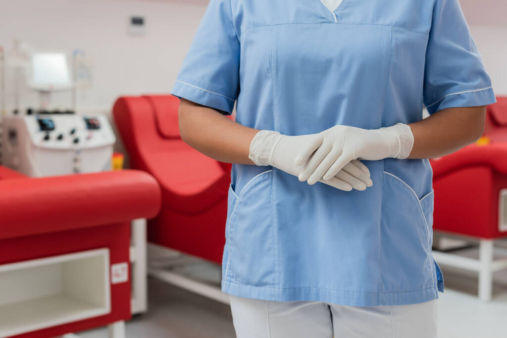 Mavi üniformalı ve lateks eldivenli çok ırklı sağlık çalışanının kan bağışı merkezinde bulanık tıbbi sandalyelerin ve kan nakli makinelerinin yanında durduğunu kısmen görmek. - Fotoğraf, Görsel