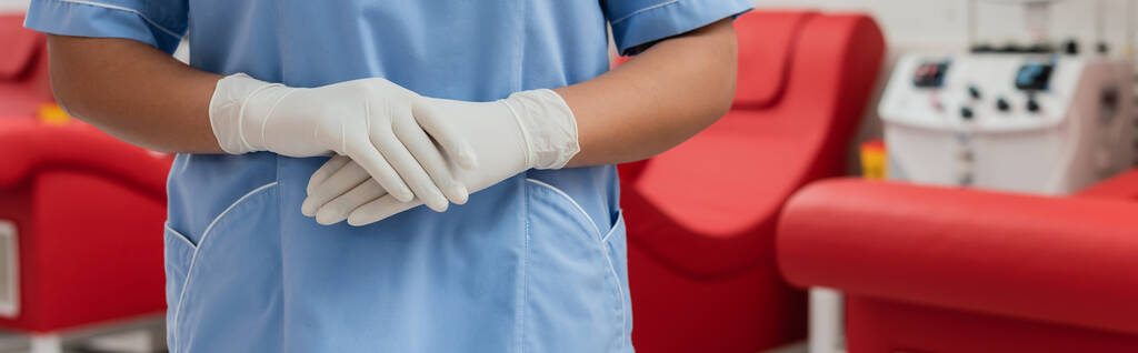 vista ritagliata di operatore sanitario multirazziale in uniforme blu e guanti in lattice in piedi in ambiente sterile del centro trasfusione di sangue, banner - Foto, immagini