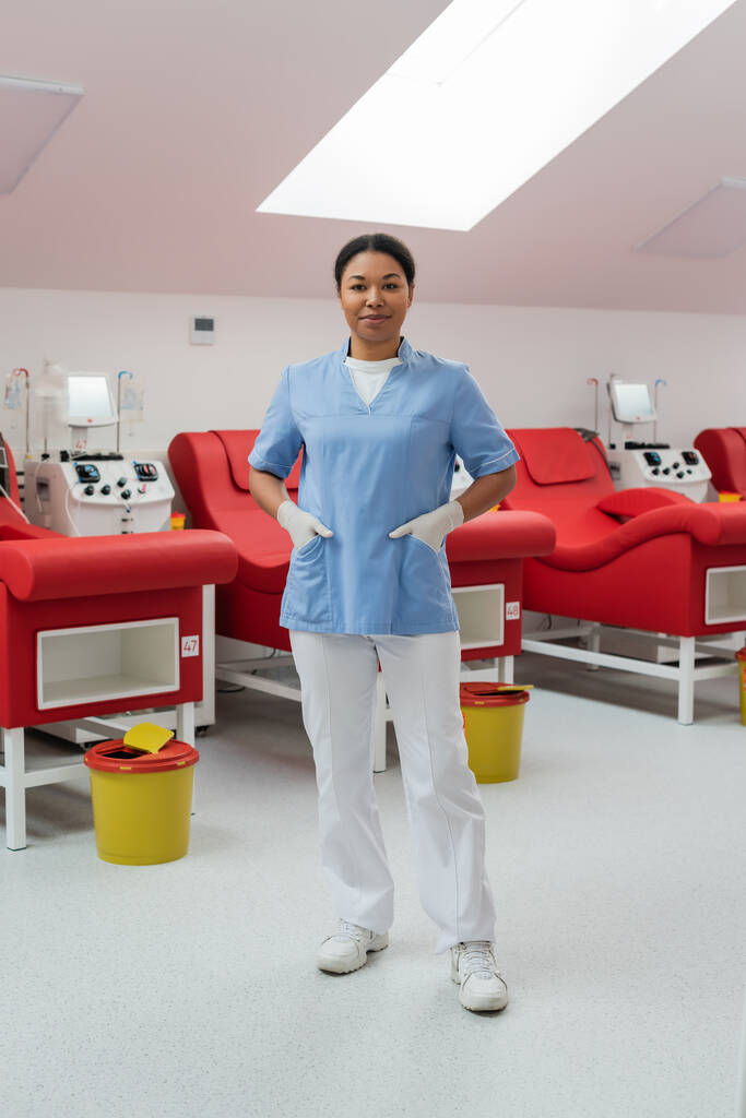 positiivinen monirotuinen sairaanhoitaja lateksikäsineissä, jotka seisovat kädet univormutaskussa lähellä lääketieteellisiä tuoleja, verensiirtokoneita ja roskakoreja verenluovutuskeskuksessa - Valokuva, kuva