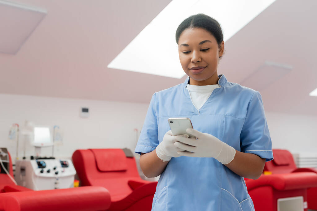 dodatni wielorasowy lekarz w niebieskim mundurze i lateksowe rękawiczki wiadomości na smartfonie w pobliżu maszyny transfuzyjnej i krzesła medyczne na rozmytym tle w centrum krwiodawstwa - Zdjęcie, obraz