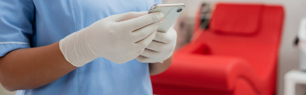 частковий вид на багаторасового медичного працівника в синій формі і латексні рукавички обміну на смартфоні поблизу розмитого медичного крісла в станції переливання крові, банер
 - Фото, зображення
