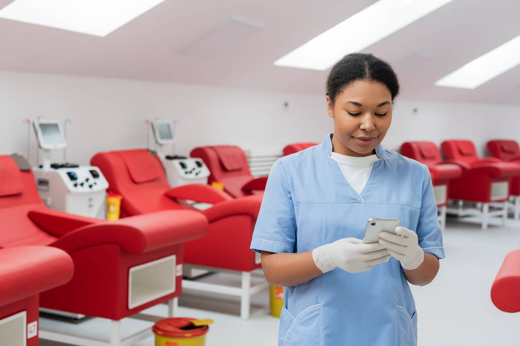 positive multiracial nurse in blue uniform and latex gloves texting on smartphone near blurred medical chairs and transfusion machines in blood donation center - Photo, Image