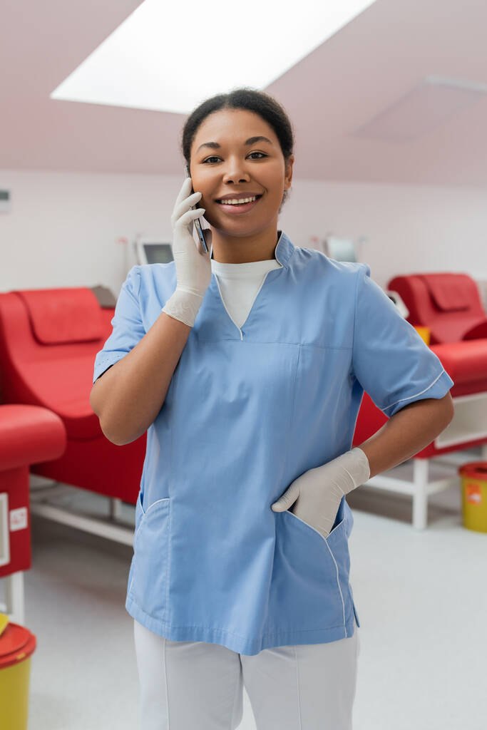 χαρούμενη πολυφυλετική νοσοκόμα σε γάντια λατέξ στέκεται με το χέρι στην τσέπη του μπλε στολή και μιλώντας σε smartphone κοντά θολή ιατρικές καρέκλες στο εργαστήριο - Φωτογραφία, εικόνα