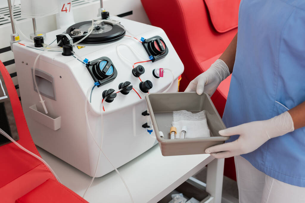 częściowy widok wielorasowego pracownika służby zdrowia w rękawiczkach mundurowych i lateksowych trzymającego tacę medyczną z probówkami w pobliżu urządzenia transfuzyjnego w centrum krwiodawstwa - Zdjęcie, obraz