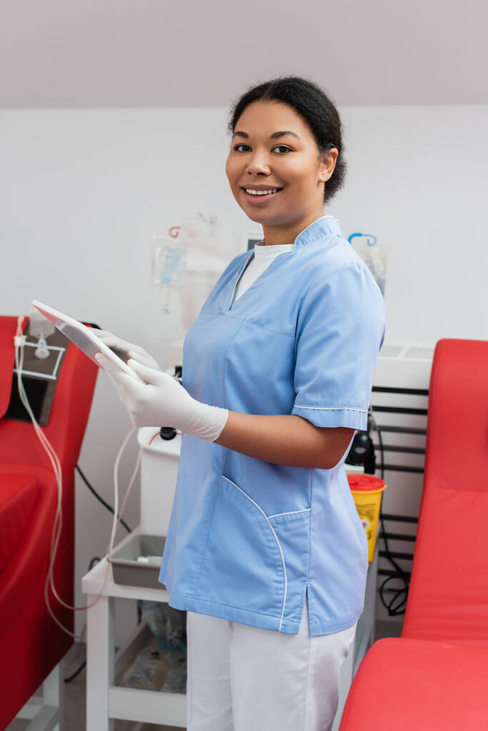 radosny wielorasowy pracownik służby zdrowia w rękawiczkach mundurowych i lateksowych trzymający tablet cyfrowy i patrzący przed kamerą w pobliżu krzeseł medycznych i automatycznej maszyny transfuzyjnej w centrum krwiodawstwa - Zdjęcie, obraz