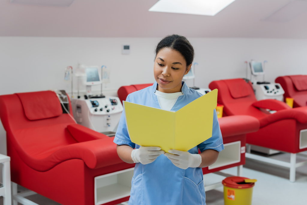 többfunkciós egészségügyi dolgozó kék egyenruhában és latex kesztyűben orvosi kartonokat olvas homályos transzfúziós gépek, orvosi székek és szemetes vödör mellett laboratóriumban - Fotó, kép