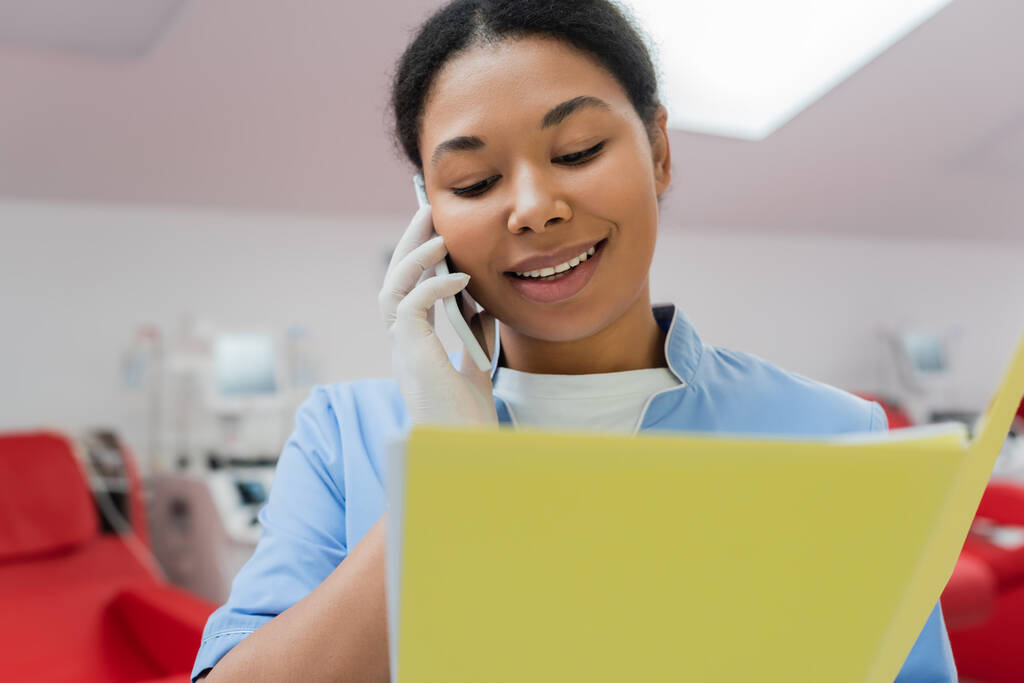 veselé multiraciální zdravotní sestra v modré uniformě a latexové rukavice mluví na smartphone a při pohledu na papírové složky v blízkosti rozmazané transfuzní stroje a lékařské židle v centru dárcovství krve - Fotografie, Obrázek