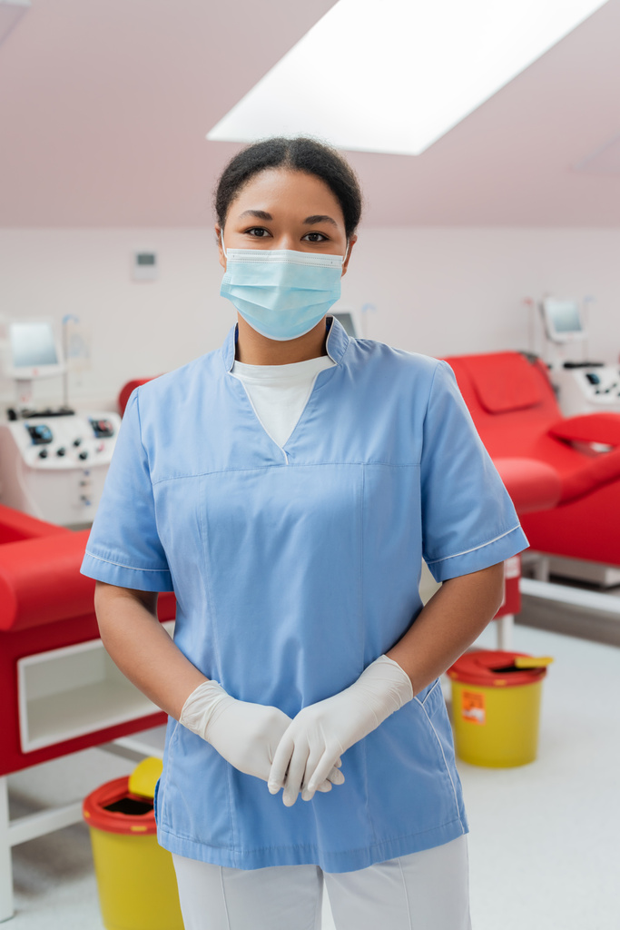 multiraciale verpleegster in blauw uniform, medisch masker en latex handschoenen kijkend naar camera in de buurt van transfusiemachines, medische stoelen en vuilnisemmers op wazige achtergrond in bloeddonatiecentrum - Foto, afbeelding