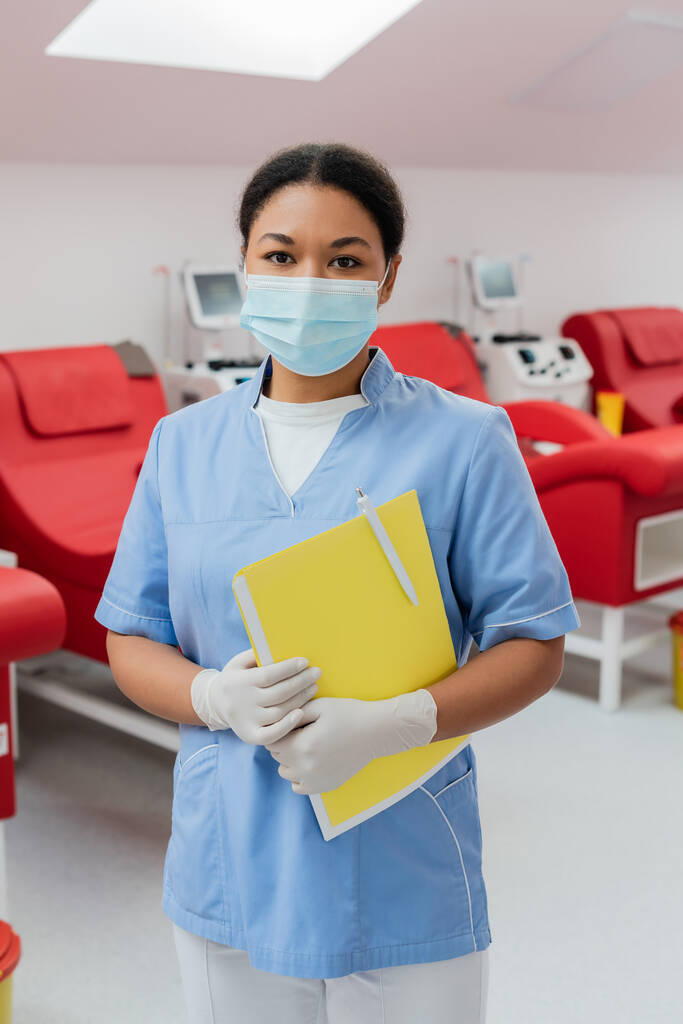 multiraciální lékař v modré uniformě, lékařská maska a latexové rukavice držící papírové složky a pero při pohledu do kamery v blízkosti rozmazaných transfuzních strojů a lékařských židlí v nemocnici  - Fotografie, Obrázek