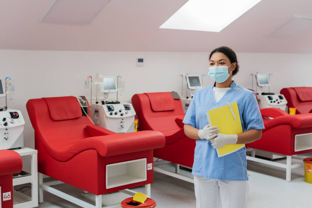 血液提供センターの人間工学に基づいた医療椅子と輸血機の近くに紙のフォルダとペンで立っている制服、医療マスクとラテックス手袋の多人種医療従事者 - 写真・画像