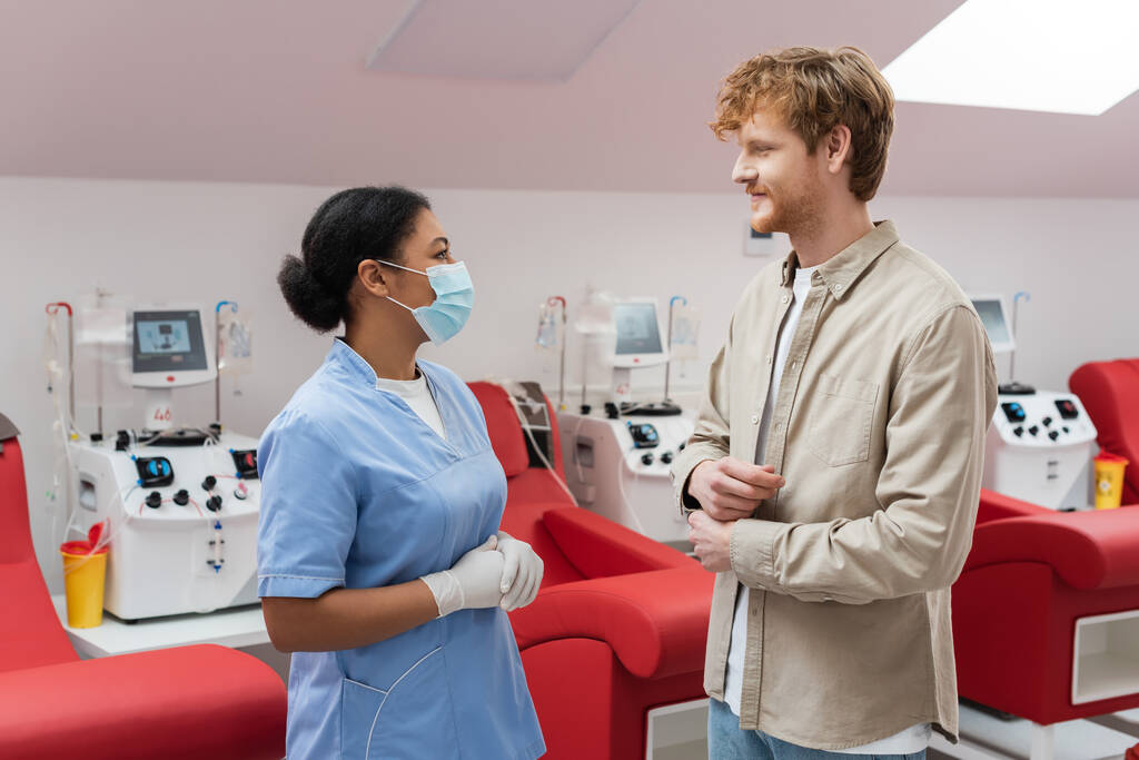 multiraciale verpleegkundige in blauw uniform, medisch masker en latex handschoenen in gesprek met roodharige vrijwilliger in de buurt van transfusiemachines en medische stoelen in het laboratorium - Foto, afbeelding