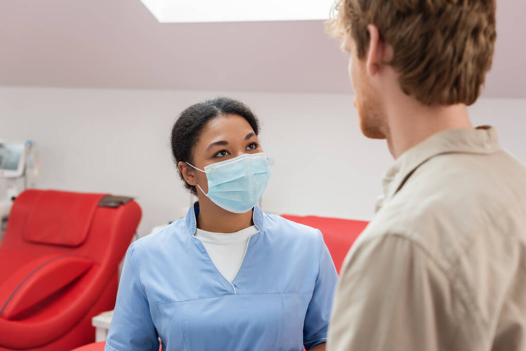 πολυφυλετικός εργαζόμενος υγειονομικής περίθαλψης με μπλε στολή και ιατρική μάσκα μιλώντας σε νεαρή κοκκινομάλλα εθελοντής σε θολή πρώτο πλάνο κοντά σε ιατρικές καρέκλες στο κέντρο αιμοδοσίας - Φωτογραφία, εικόνα