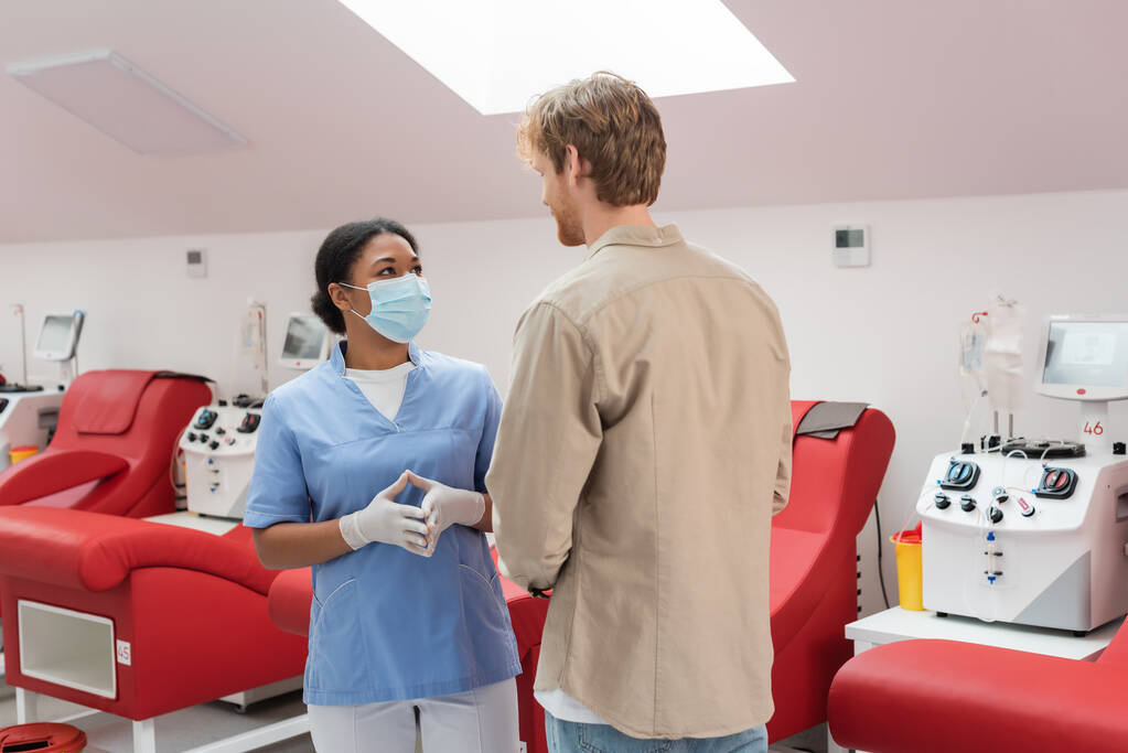 wielorasowy pracownik służby zdrowia w mundurze, masce medycznej i rękawiczkach lateksowych rozmawiający z rudowłosym mężczyzną w pobliżu krzeseł medycznych i automatów transfuzyjnych w klinice - Zdjęcie, obraz
