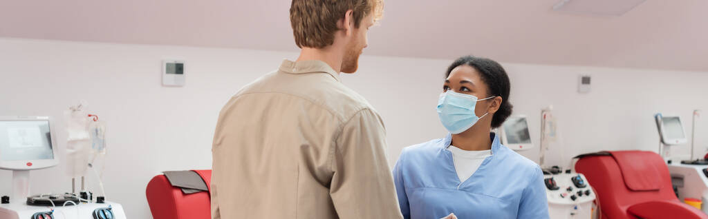 mladá zrzka dárce krve a multiraciální zdravotní sestra v modré uniformě a lékařská maska mluví v blízkosti lékařských židlí a transfuzních strojů v nemocnici, banner - Fotografie, Obrázek