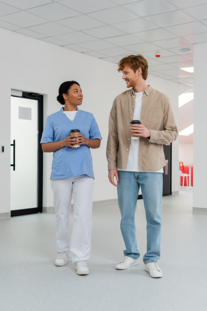 multiraciale verpleegster in uniform en jong, positieve roodharige vrijwilliger met papieren bekers met koffie en praten in de wachtkamer van het bloeddonatiecentrum, deur op de achtergrond  - Foto, afbeelding