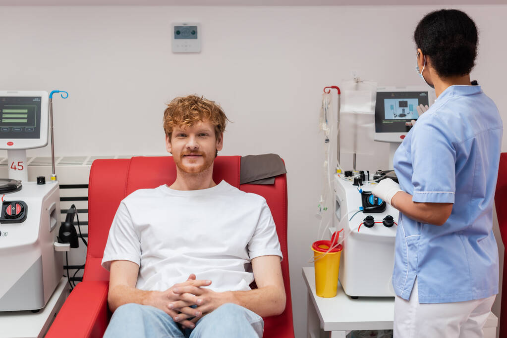 wielorasowa pielęgniarka w niebieskim mundurze operująca maszyną transfuzyjną w pobliżu plastikowej filiżanki i rudowłosej wolontariuszki siedzącej na krześle medycznym i patrzącej przed kamerą w centrum krwiodawstwa - Zdjęcie, obraz
