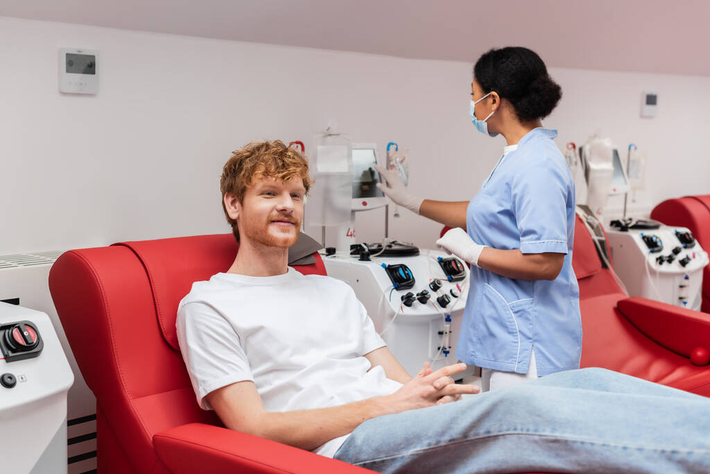 πολυφυλετική νοσοκόμα στην ιατρική μάσκα και λατέξ γάντια που λειτουργούν μετάγγιση μηχάνημα σχεδόν ευχαριστημένος κοκκινομάλλα άνθρωπος κάθεται σε άνετη καρέκλα στο κέντρο αιμοδοσίας - Φωτογραφία, εικόνα