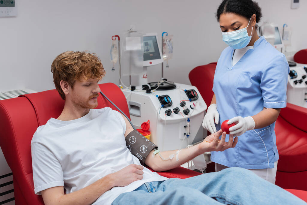 többfunkciós egészségügyi dolgozó orvosi maszkban és latex kesztyűben, gumilabdát ad a vöröshajú vérnyomásmérős férfinak, aki orvosi széken ül a transzfúziós gép közelében a kórházban. - Fotó, kép