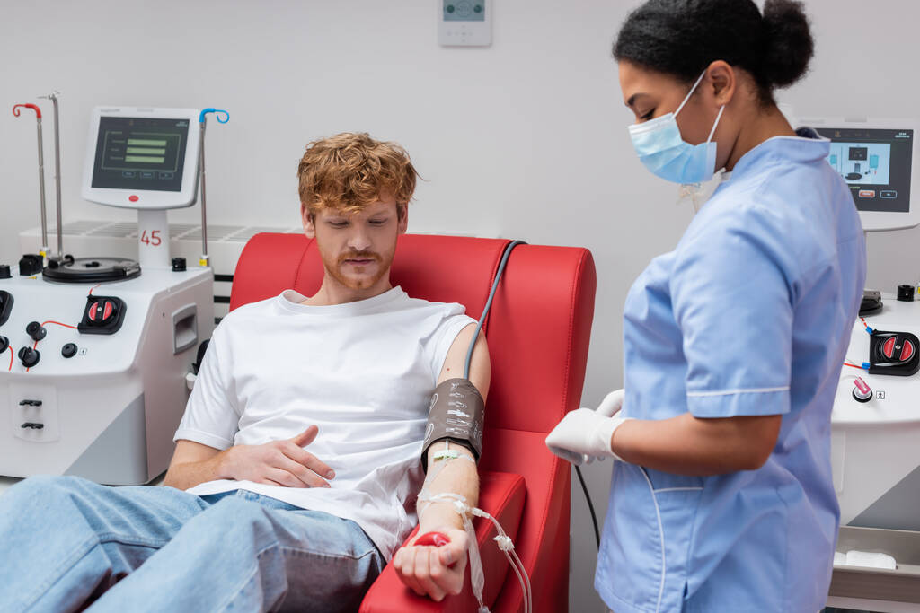 punapää vapaaehtoinen verensiirrolla asettaa puristamalla kumipallo istuessaan lääketieteellisellä tuolilla lähellä automaattisia laitteita ja monirotuinen sairaanhoitaja lääketieteellisessä naamiossa verenluovutuskeskuksessa - Valokuva, kuva