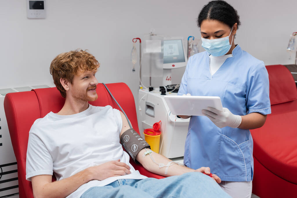 multiracial nurse in medical mask and latex gloves using digital tablet near transfusion machine and redhead donor with blood pressure cuff smiling on medical chair in laboratory - Photo, Image