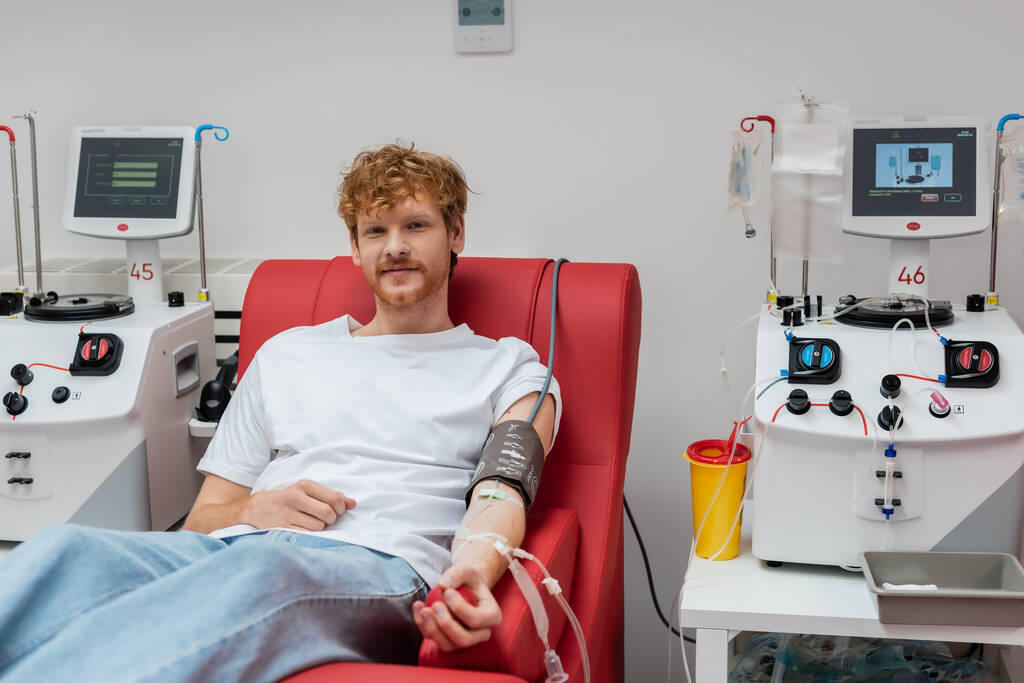 улыбающийся рыжий мужчина в наборе для переливания крови и манжеты кровяного давления сидя на медицинском стуле и глядя на камеру рядом с автоматизированным оборудованием и пластиковой чашкой в клинике - Фото, изображение