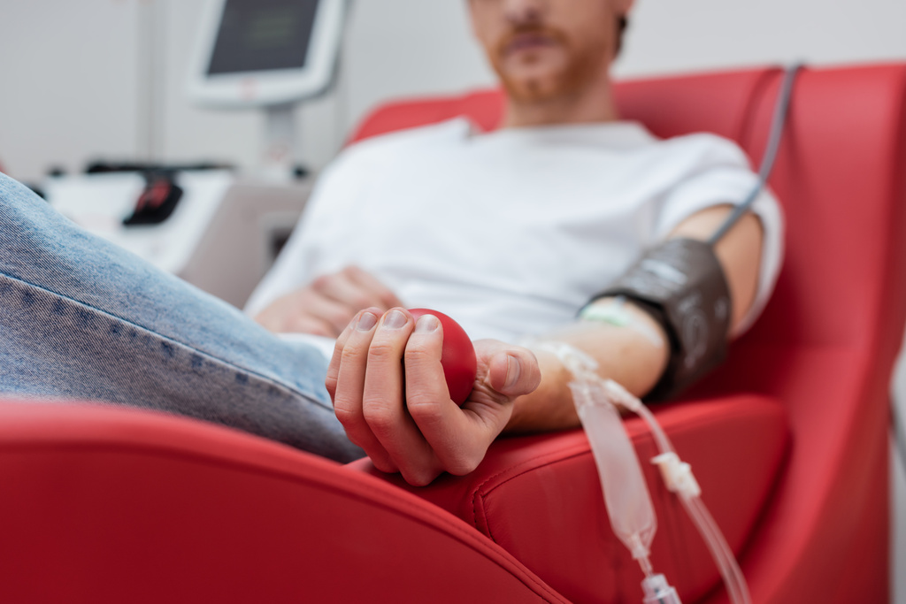 enfoque selectivo de la bola de goma en la mano de voluntario recortado con transfusión conjunto sentado en la silla médica cómoda cerca de la máquina de transfusión en el centro de donación de sangre - Foto, Imagen