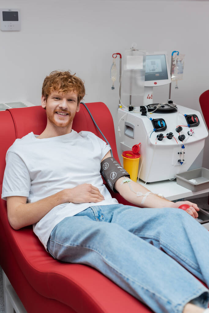 junger, fröhlicher Rotschopf mit Transfusionsset sitzt auf ergonomischem Medizinstuhl und lächelt in die Kamera neben modernen automatisierten Geräten und Plastikbechern - Foto, Bild