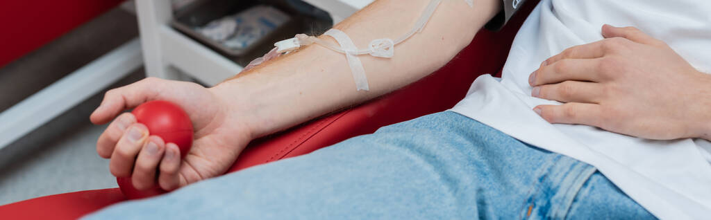 vue recadrée du volontaire avec ensemble de transfusion serrant la balle en caoutchouc tout en étant assis sur une chaise médicale confortable dans le centre moderne de don de sang, bannière - Photo, image