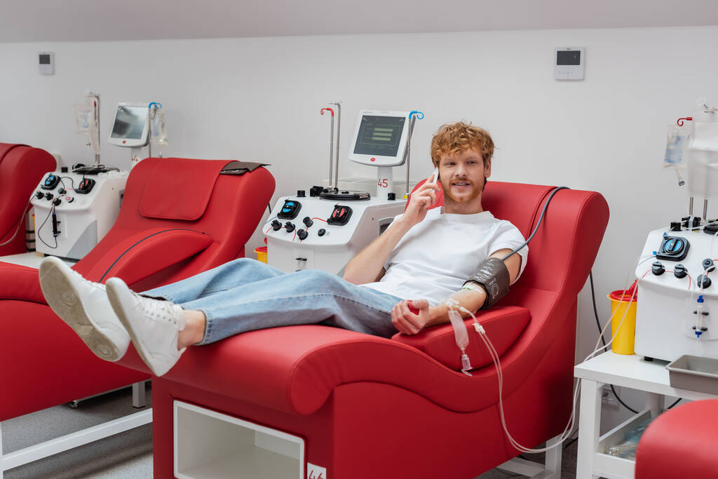 junger rothaariger Mann mit Blutdruckmanschette sitzt auf dem Behandlungsstuhl in der Nähe von Transfusionsmaschinen und Plastikbecher, während er während der Blutspende in der Klinik mit dem Handy telefoniert - Foto, Bild