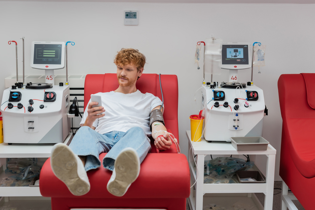 молодой рыжий волонтер сидит на эргономичном медицинском стуле и отправляет сообщения на смартфон во время процедуры сдачи крови возле автоматизированного оборудования и пластиковой чашки в клинике - Фото, изображение