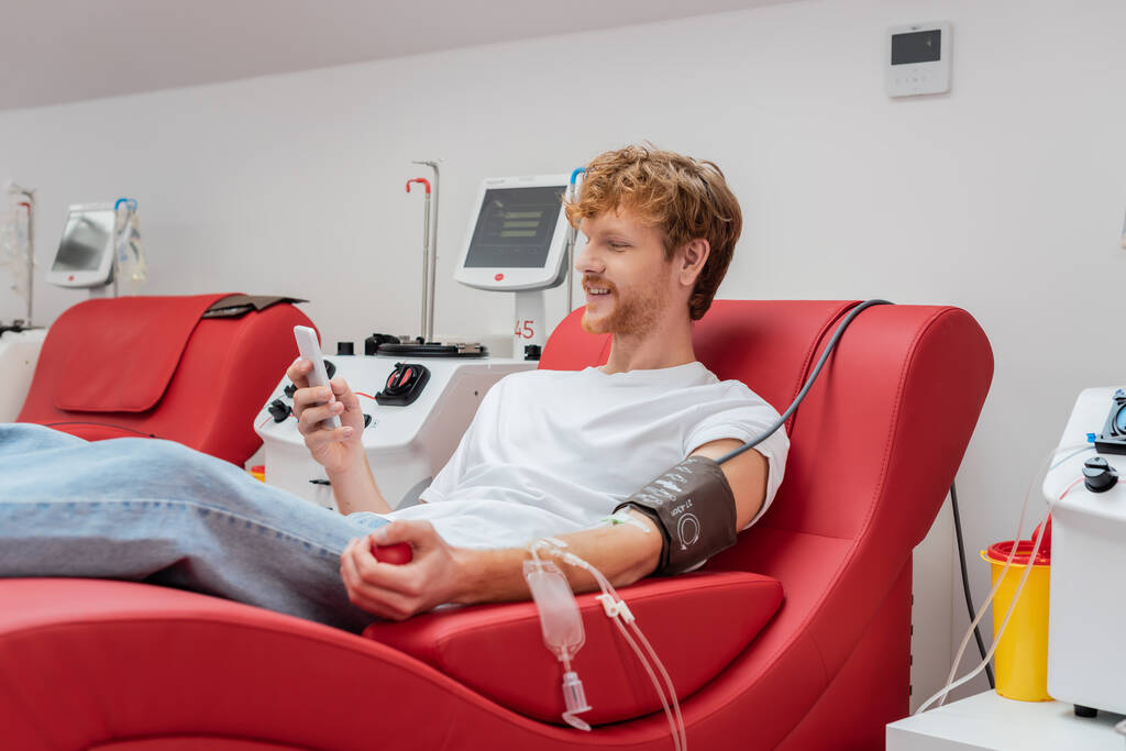 ruda i uśmiechnięty człowiek przeglądający internet na telefon komórkowy w fotelu medycznym w pobliżu automatów transfuzyjnych i plastikowych kubków w centrum krwiodawstwa - Zdjęcie, obraz