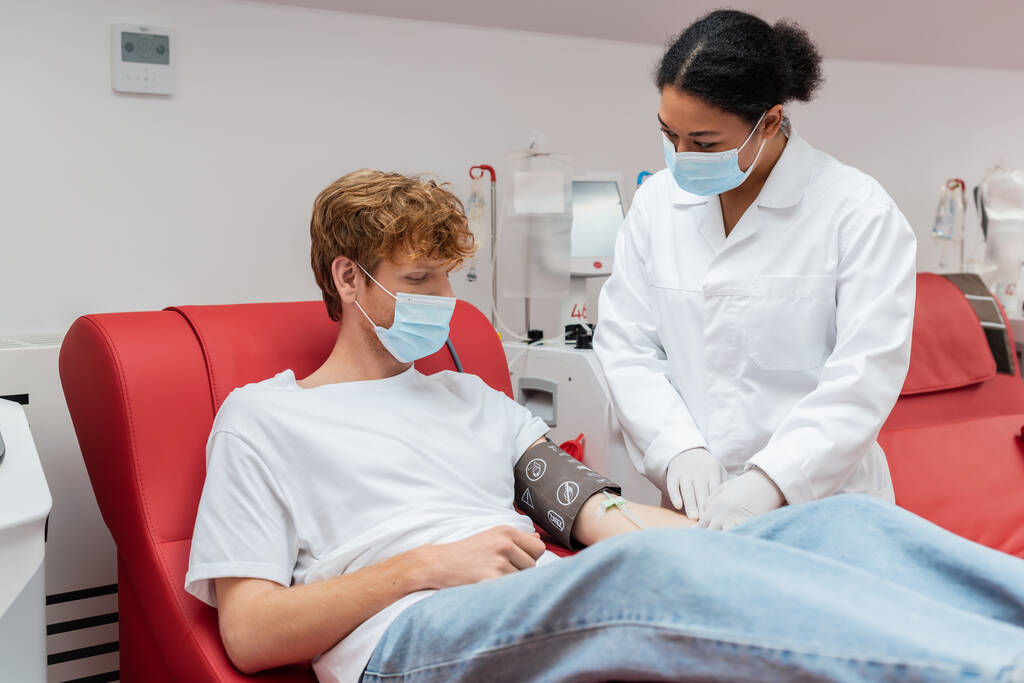medico multirazziale in camice bianco e maschera medica guardando volontario rossa con polsino di pressione sanguigna seduto vicino alle attrezzature automatizzate nel centro di donazione di sangue - Foto, immagini