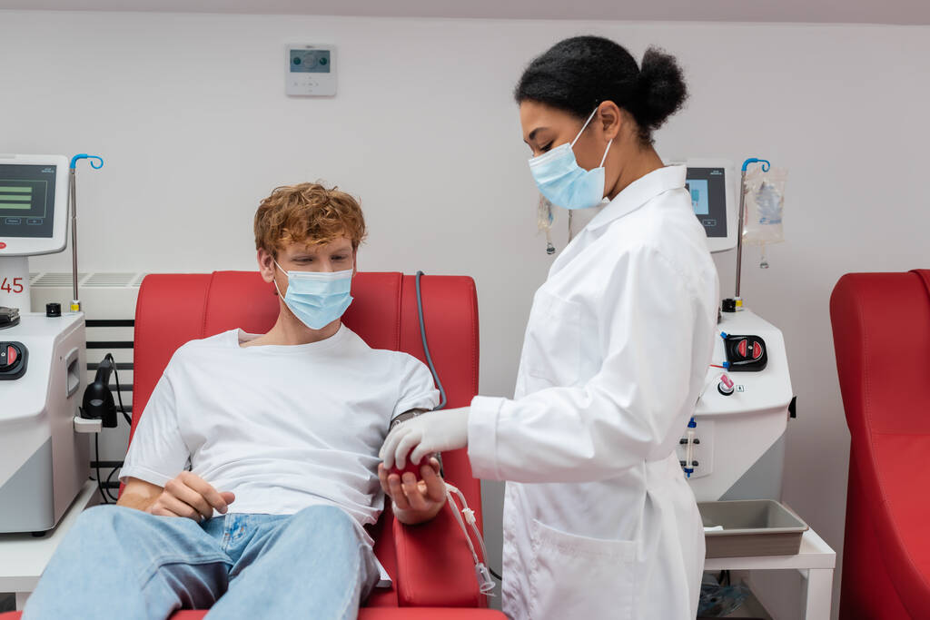 médecin multiracial en masque médical et manteau blanc donnant balle en caoutchouc à un volontaire roux assis sur une chaise ergonomique près des machines à transfusion dans un centre de don de sang - Photo, image