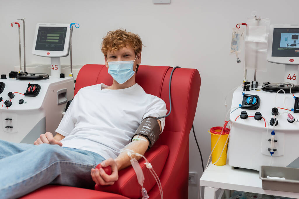 redhead volunteer in medical mask and blood pressure cuff looking at camera on comfortable chair near automated transfusion machines in modern laboratory - Photo, Image