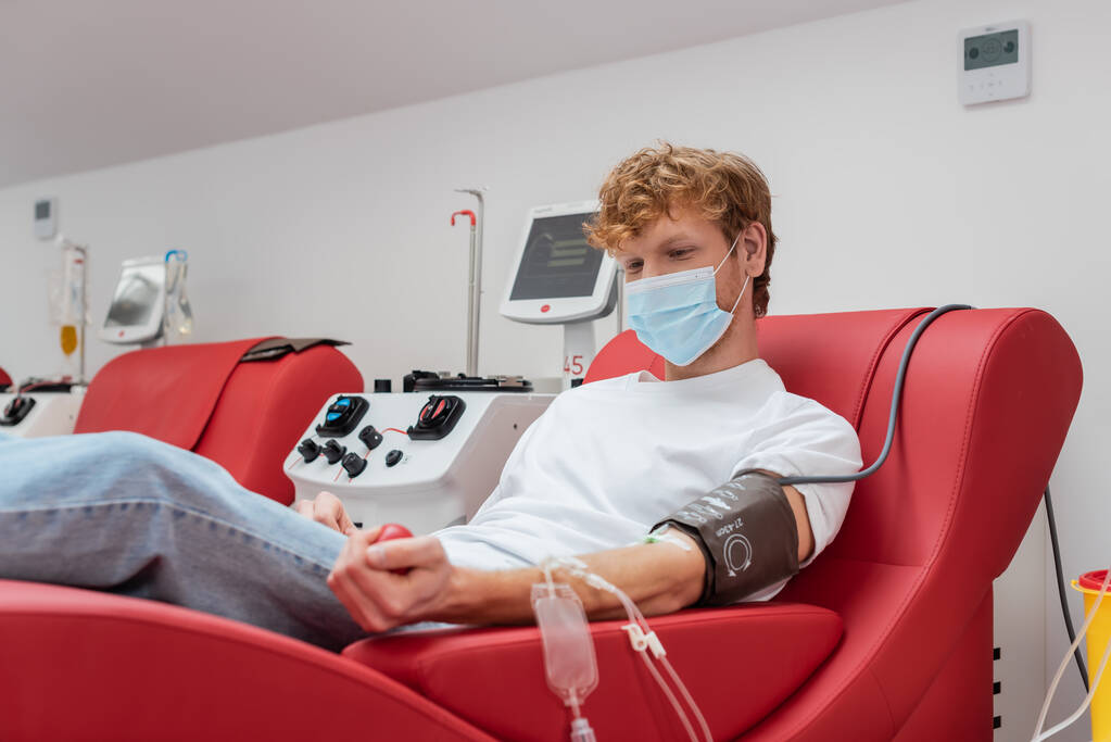 Молодой рыжеволосый донор в медицинской маске и при переливании крови сжимает резиновый мячик, сидя на удобном стуле рядом с автоматизированным оборудованием во время процедуры сдачи крови в клинике - Фото, изображение