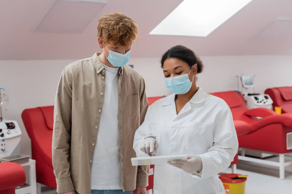 multiracial doctor in medical mask pointing at digital tablet near redhead man and medical chairs with transfusion machines in blood donation center - Photo, Image