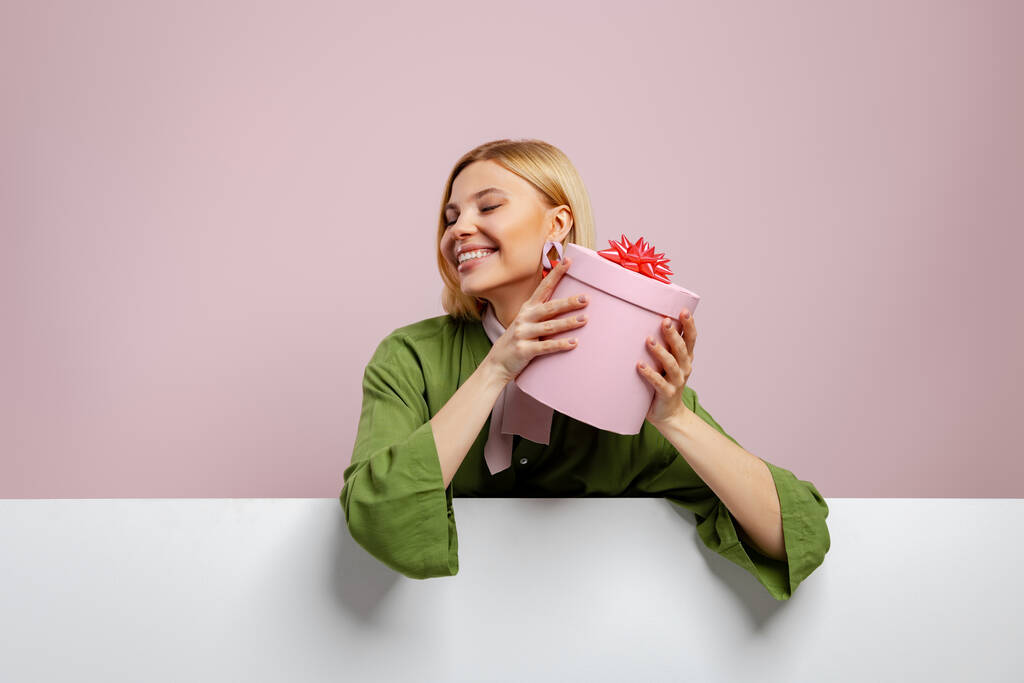 Çekici genç bir kadın hediye kutusunu yüzüne yakın tutuyor ve pembe arka planda gülümsüyor. - Fotoğraf, Görsel