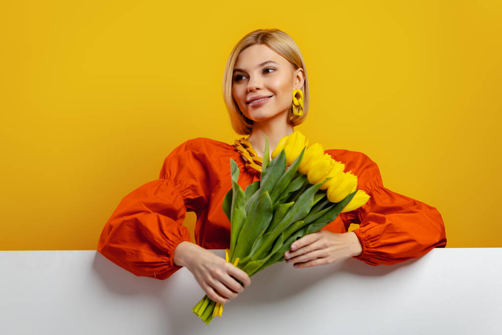 Atrakcyjna młoda kobieta trzyma garść tulipanów i uśmiecha się na żółtym tle - Zdjęcie, obraz