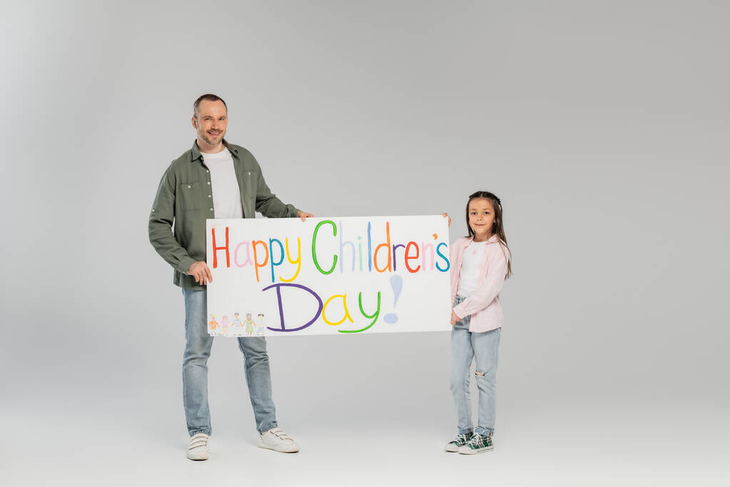 Volledige lengte van de glimlachende vader en tiener dochter in casual kleding kijken naar de camera terwijl het vasthouden van bordje met gelukkige kinderdag belettering terwijl samen vieren op grijze achtergrond - Foto, afbeelding