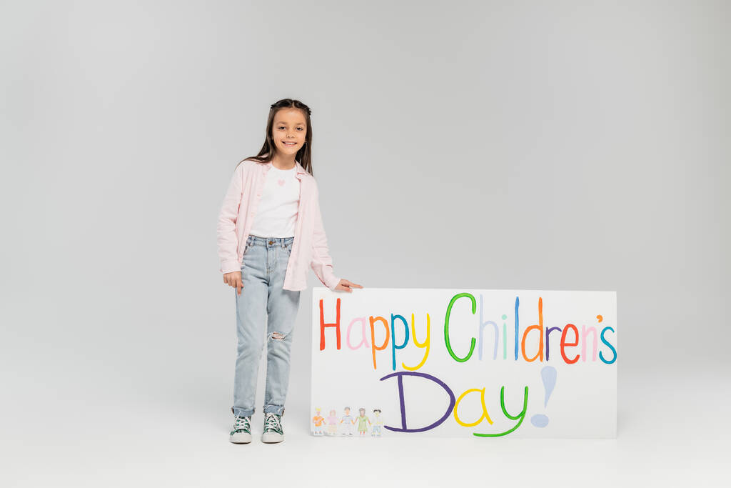 Улыбающаяся девочка-подросток в повседневной одежде смотрит в камеру, стоя рядом с плакатом с надписью "Счастливый детский день" во время отдыха в июне на сером фоне с копировальным пространством  - Фото, изображение