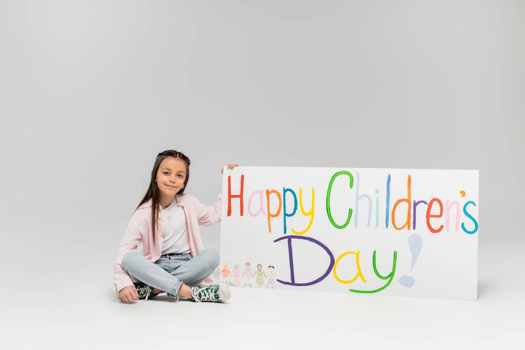 Glimlachend meisje in casual kleding kijkend naar de camera tijdens het zitten in de buurt van bordje met gelukkige kinderdag belettering tijdens vakantie in juni op grijze achtergrond - Foto, afbeelding
